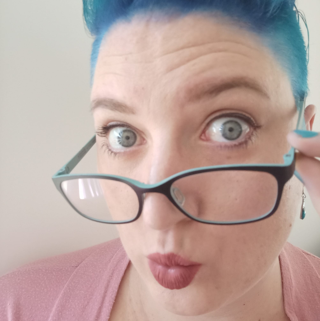 a woman with blue hair and blue eyes looking over her glasses while wearing mauve lipstick and a pink shirt