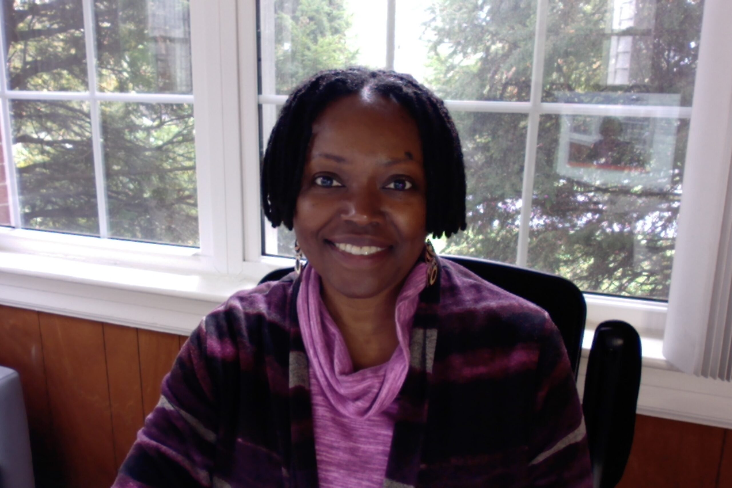 a Black woman smiling at the camera while wearing a purple and maroon striped cardigan and purple shirt