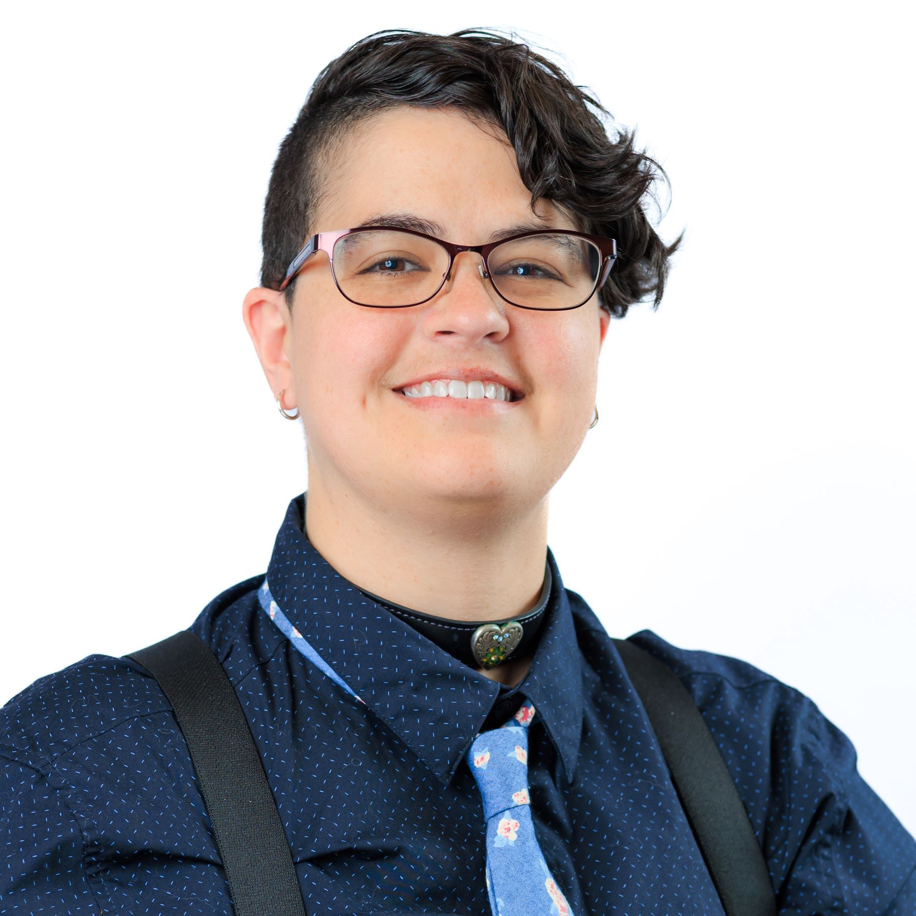 a Caucasian person with black hair and wearing glasses, a blue dress shirt, and a blue tie smiles at the camera