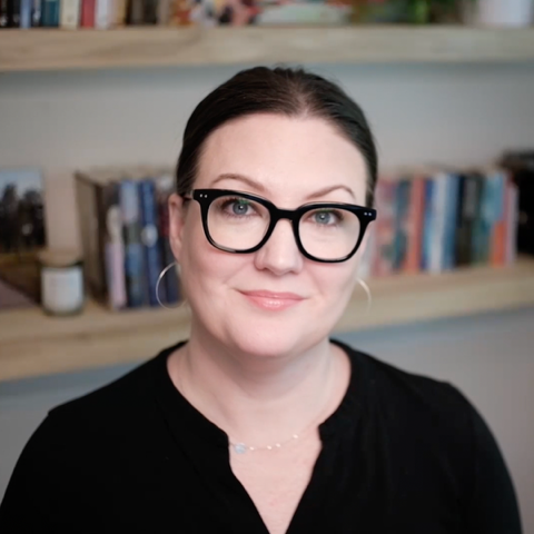 a smiling Caucasian woman with dark hair and wearing black glasses and a black shirt sitting in front of a bookshelf