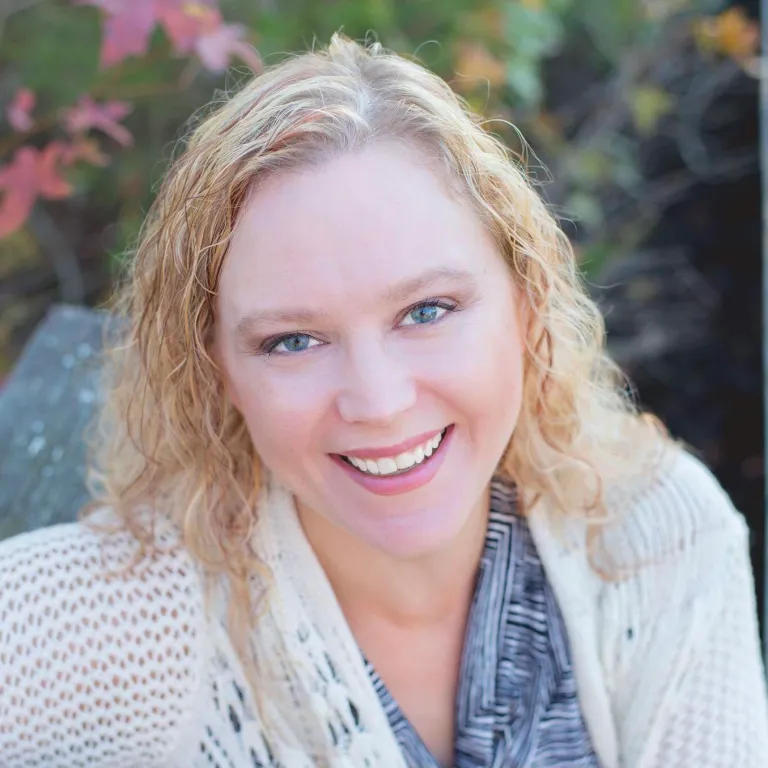 a smiling Caucasian woman with blonde hair and blue eyes wearing a white cardigan over a blue-and-white striped shirt