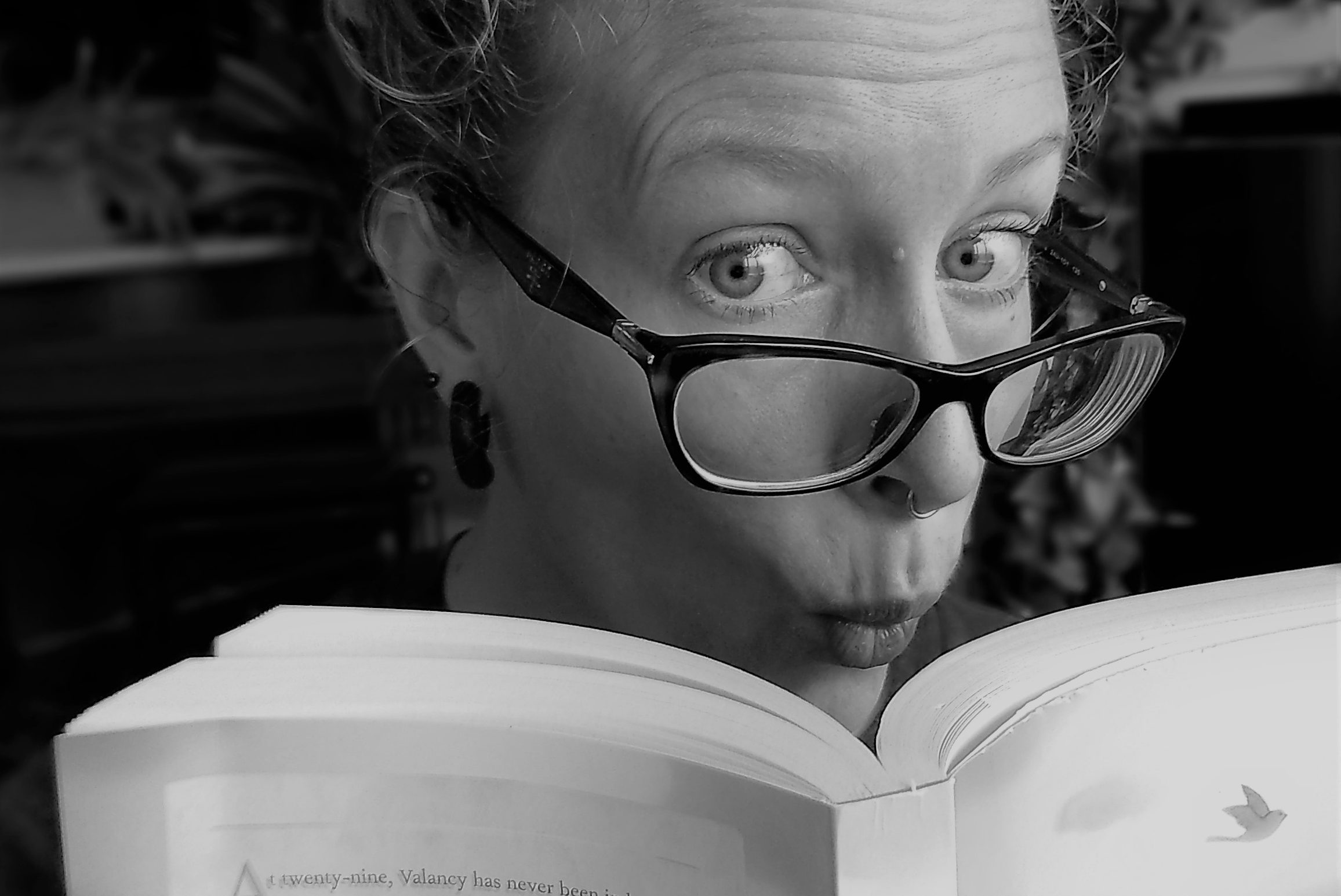 a black and white photo of a woman with black glasses looking over the top of a book, making a silly face