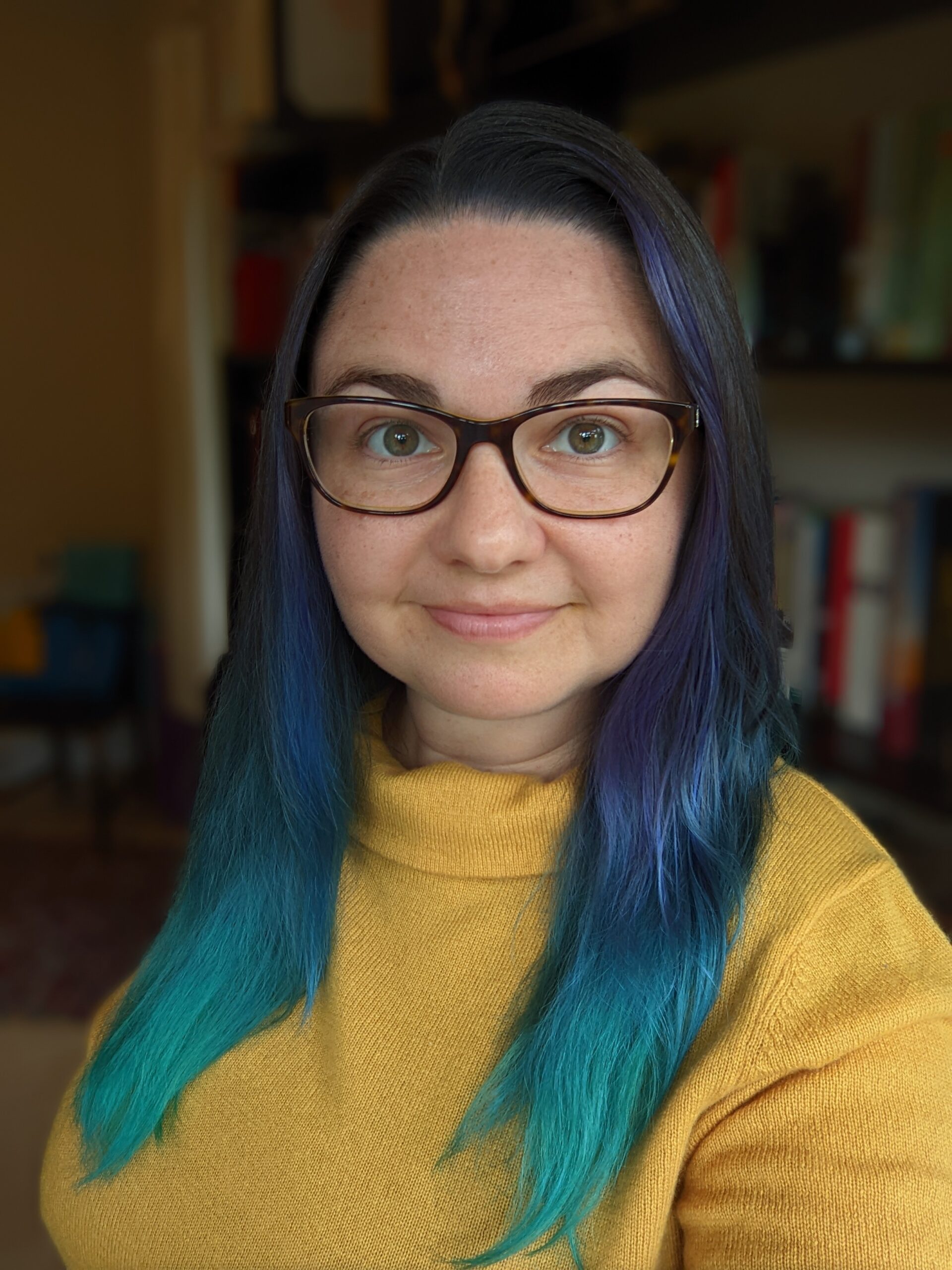 a smiling caucasian woman with dark blue hair that fades to teal at the tips wearing a yellow turtleneck and dark frame glasses