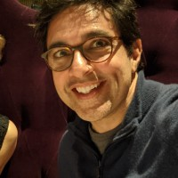 a smiling dark-skinned man with dark hair and black glasses wearing a black shirt