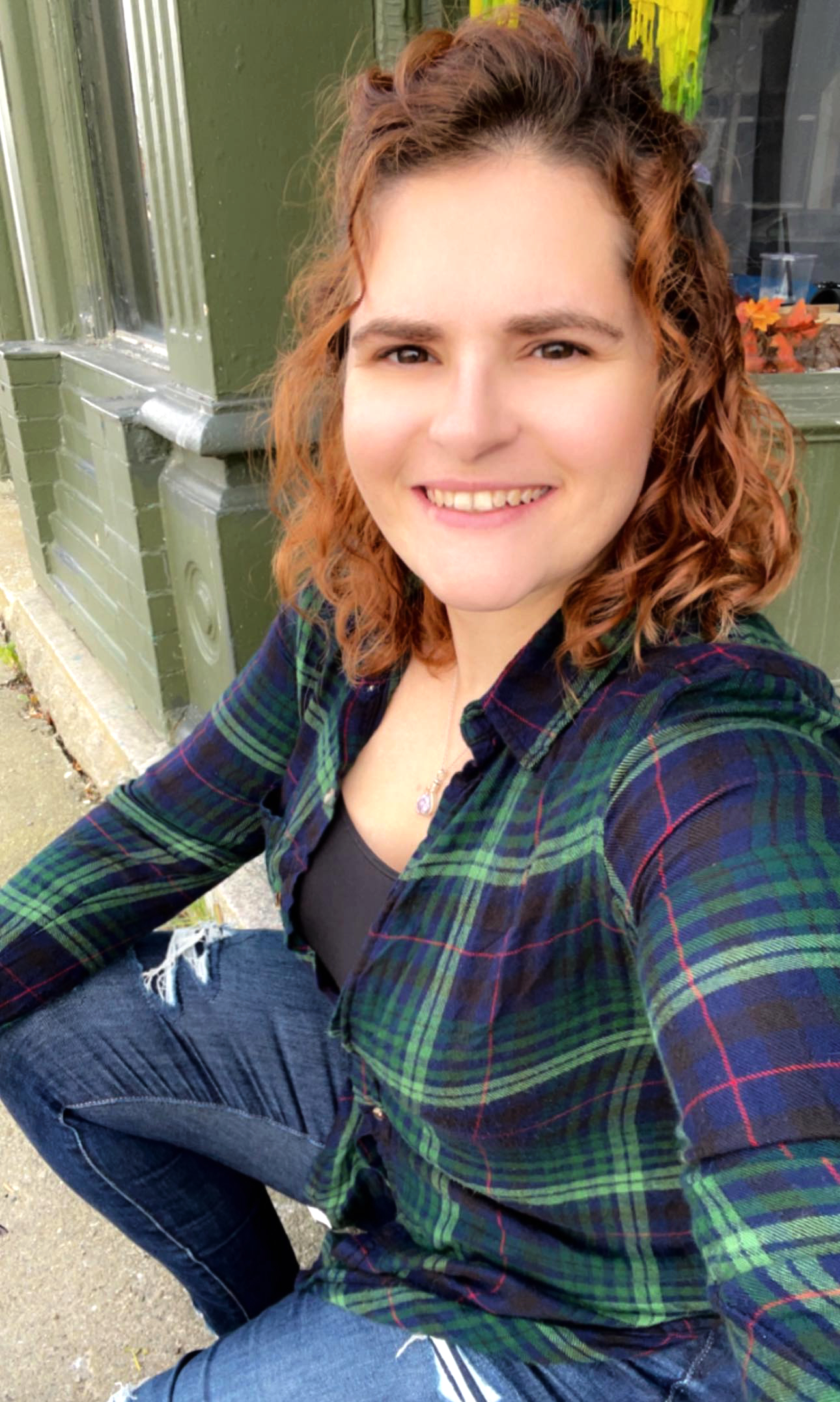 A smiling white woman with auburn hair and dark eyes wearing a blue and green flannel shirt.