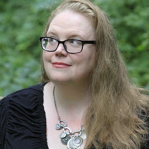 A white woman wearing black glasses looking off to her left with a whimsical smile.